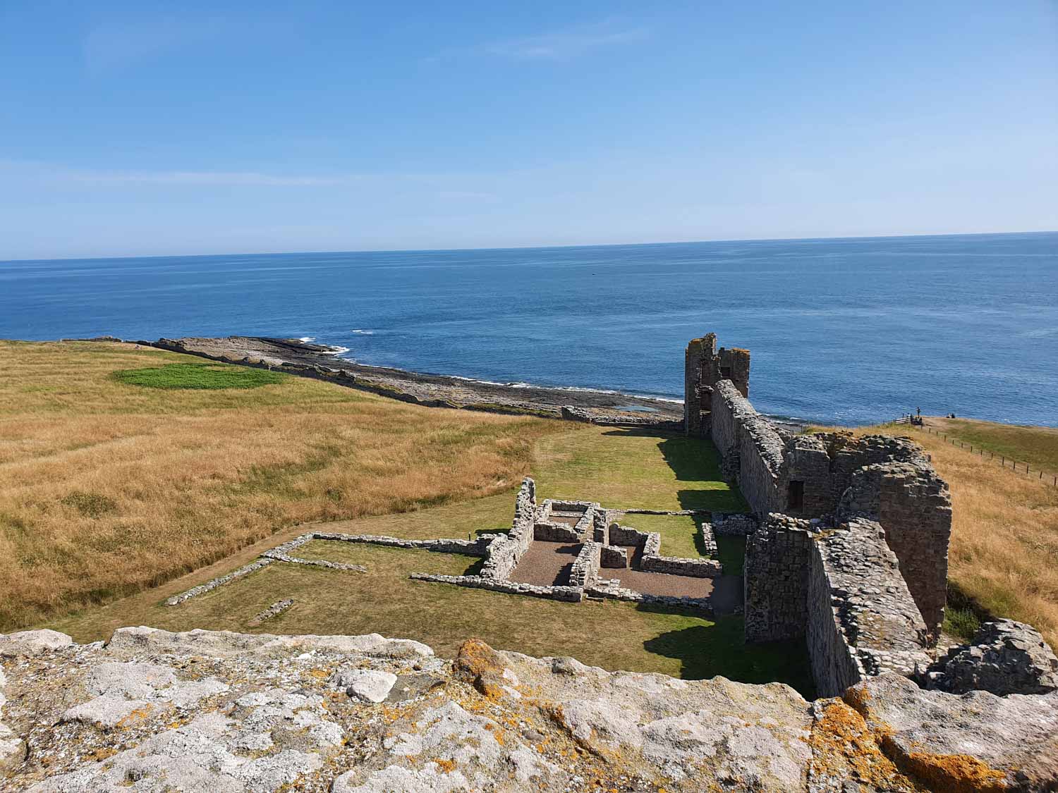 View from one of the towers down to the grey stone walls, foundations of buildings, grassy court and sea at Dunstansburgh Castle, Northumberland