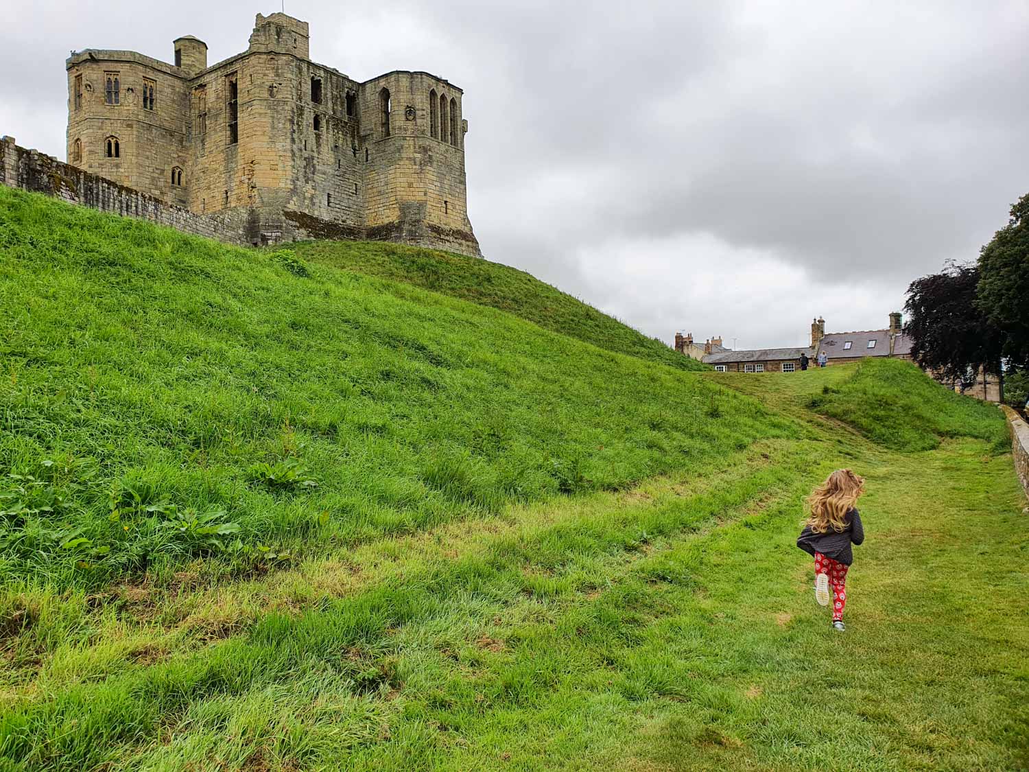My daughter runs around the moat outside Warkworth Castle Northumberland with some of the ruined stone buildings seen on the slope above - our visit to Warkworth Castle with kids