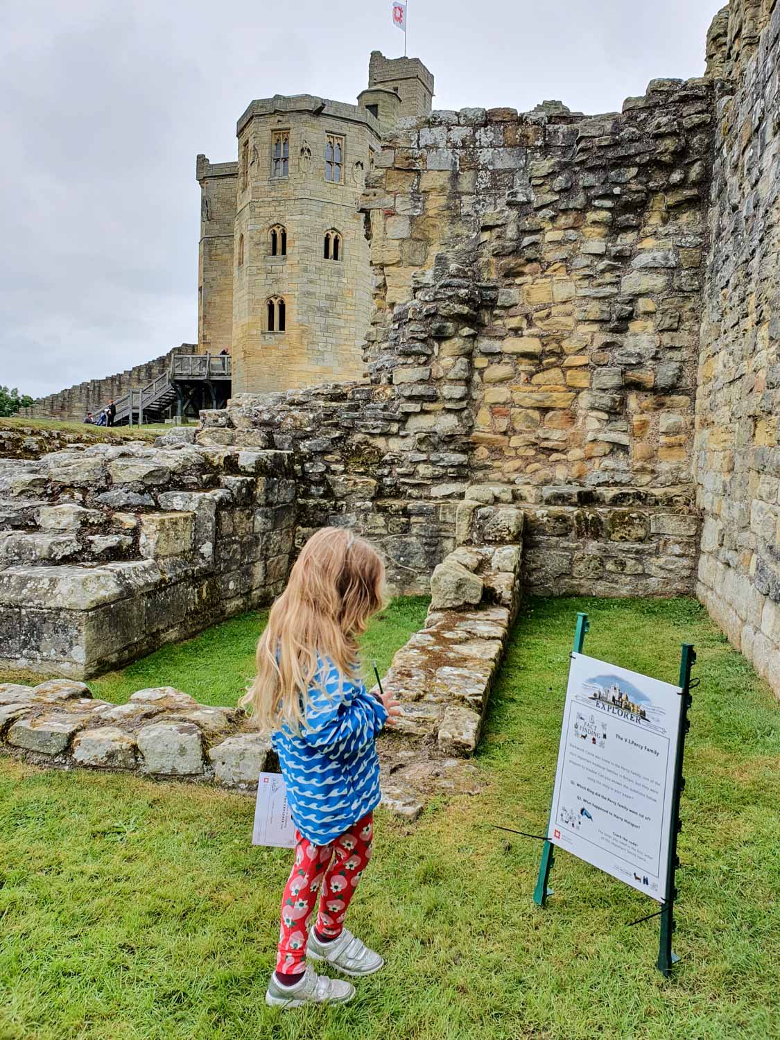 My nine-year-old daughter looks at one of the signs on the English Heritage train at Warkworth Castle, Northumberland with the ruins of the castle behind her during a day out at Warkworth Castle with kids
