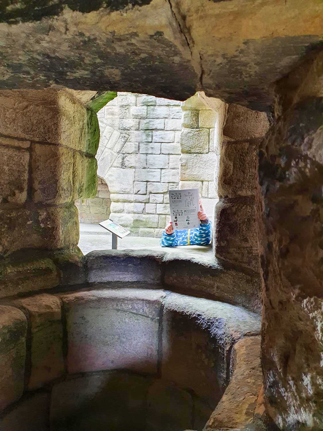Looking through stone windows to my daughter holding an Engish Heritage trail, with a huge round hole in between - visiting Warkworth Castle, Northumberland with kids