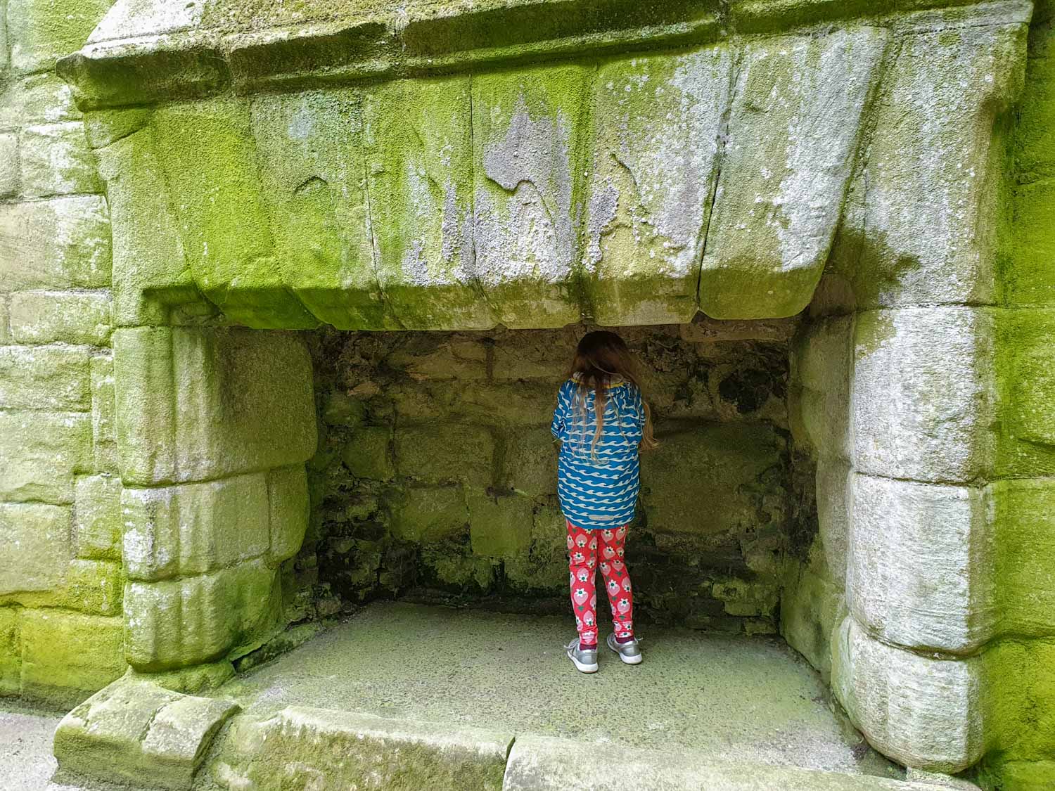 My 9-year-old daughter stands in one of the giant hearts at Warkworth Castle, with green moss on the stones - visiting Warkworth Castle with kids
