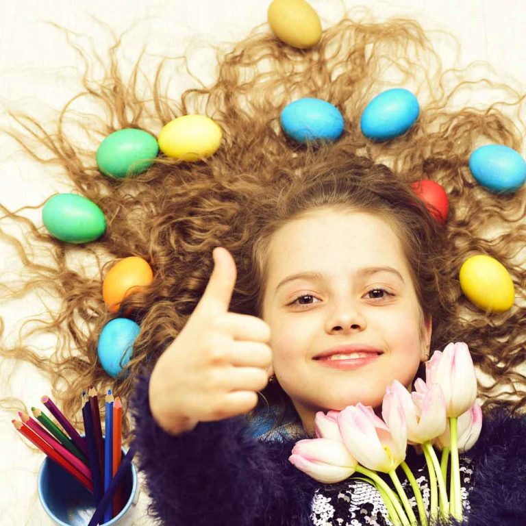 Girl lying down with bunch of tulips and her thumbs up, with brightly coloured Easter eggs in her hair - the best UK Easter days out with kids