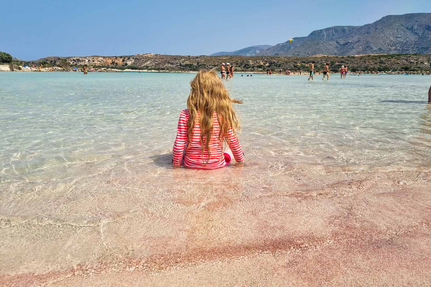 Close up of the pink sand on Elafonisi beach in Crete with my daughter sitting in the shallow clear water of the lagoon my tips for visiting Elafonisi Beach with kids