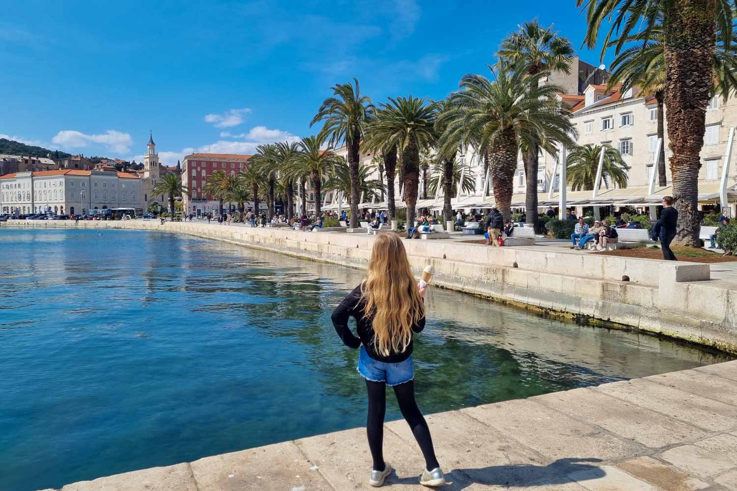 My daughter holding an ice cream looking along the Riva waterfront promenade lined with palm trees to some of the old buildings of Split - my pick of the best things to do in Split with kids