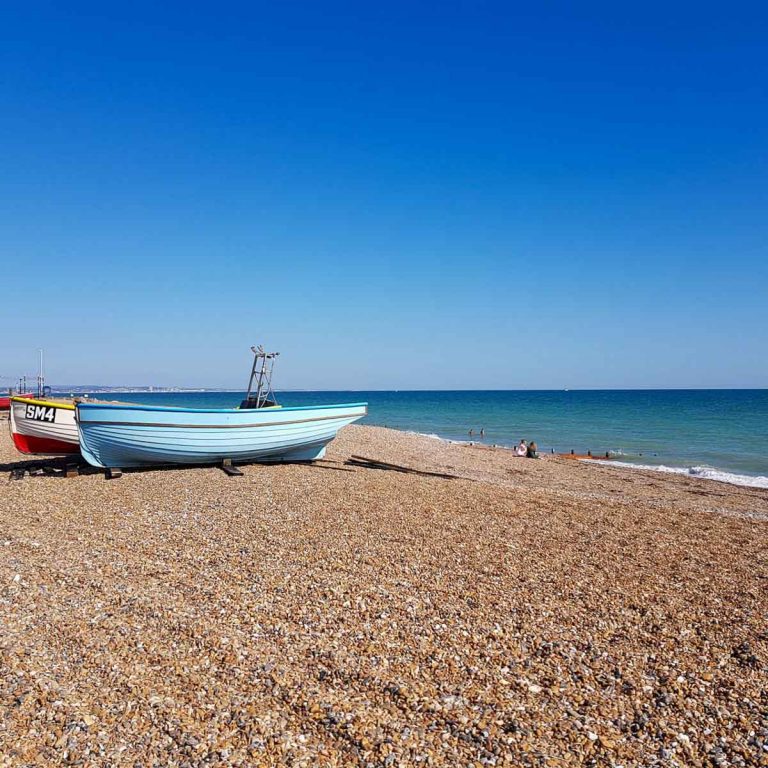 Colourful blue, red and yellow boats pulled up onto a pebble beach by the sea in West Sussex, against a blue sky - my pick of the best things to do in Sussex with kids