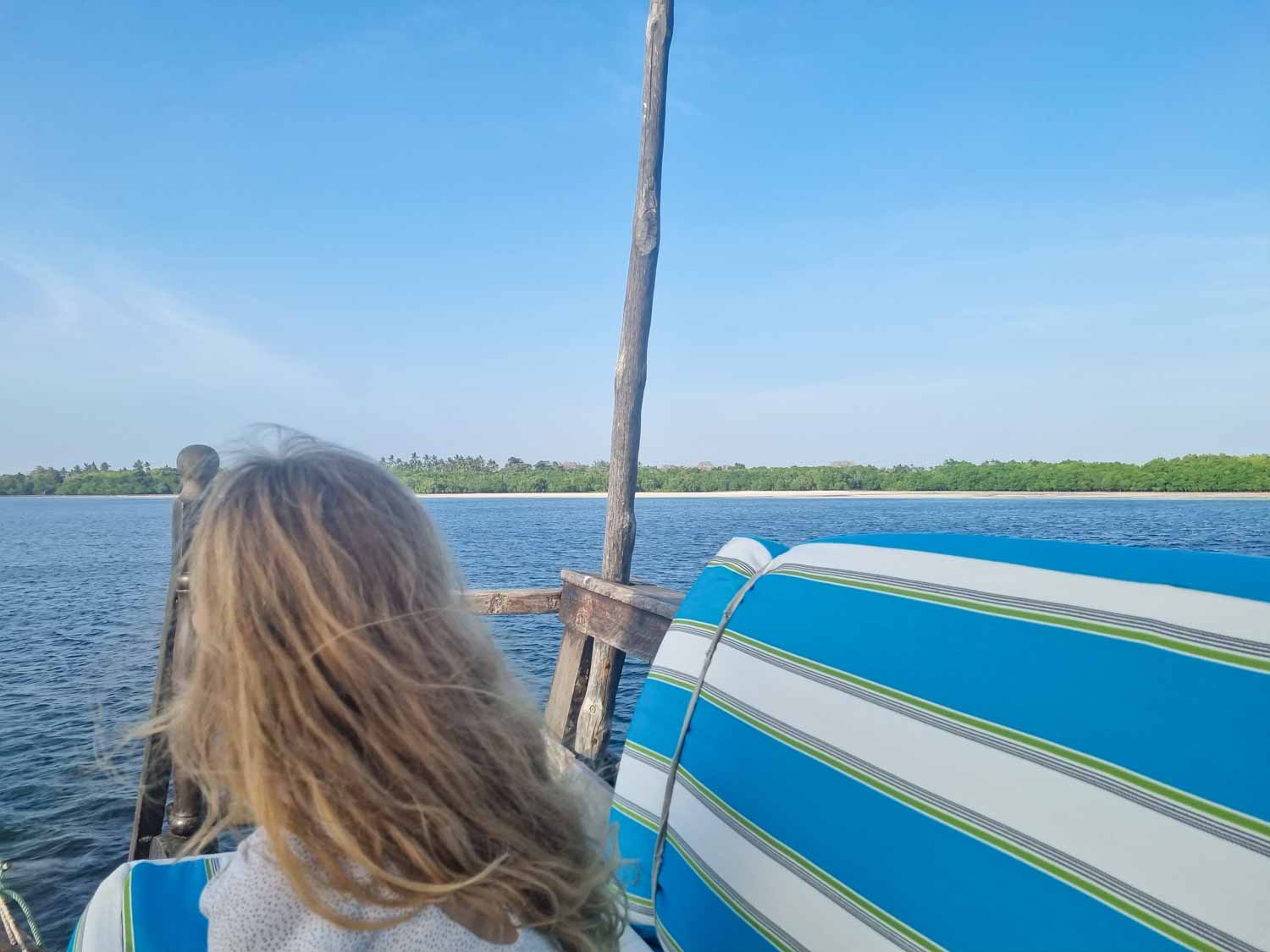 My daughter on a striped cushioned seat on the top deck of a dhow, looking out to the mangroves lining the creek during our dhow cruise arranged during a stay at Hemingways Watamu with kids