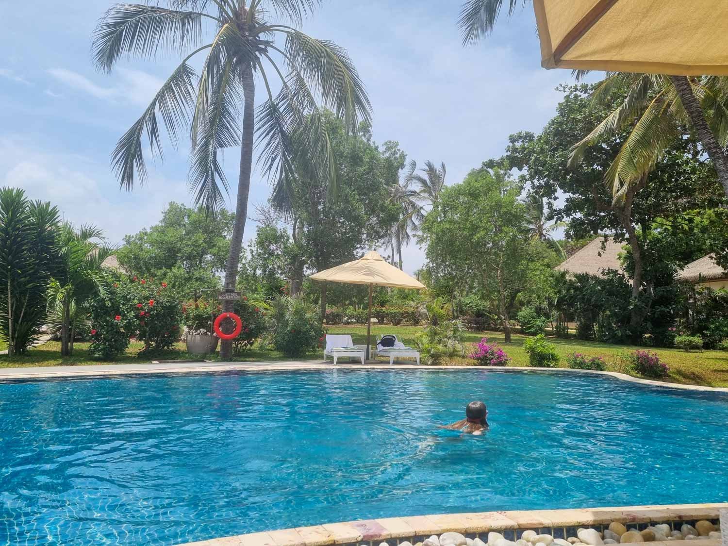 My daughter swims in the quiet pool at Hemingways Watamu in Kenya, with loungers and lush trees seen in the background - my review of Hemingways Watamu with kids