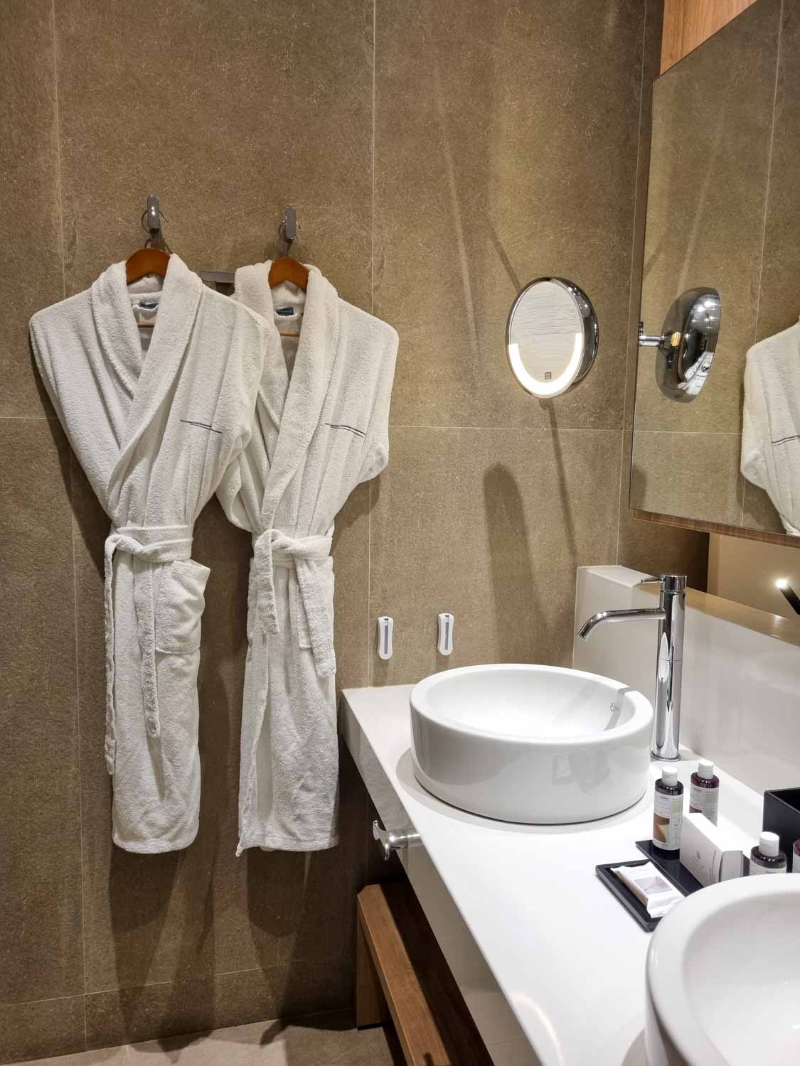 Two bathrobes hanging on the wall in the bathroom of a Junior suite at Gennadi Grand resort, with the two round sinks and toiletries just seen - my Gennadi Grand resort review of a family holiday in Rhodes