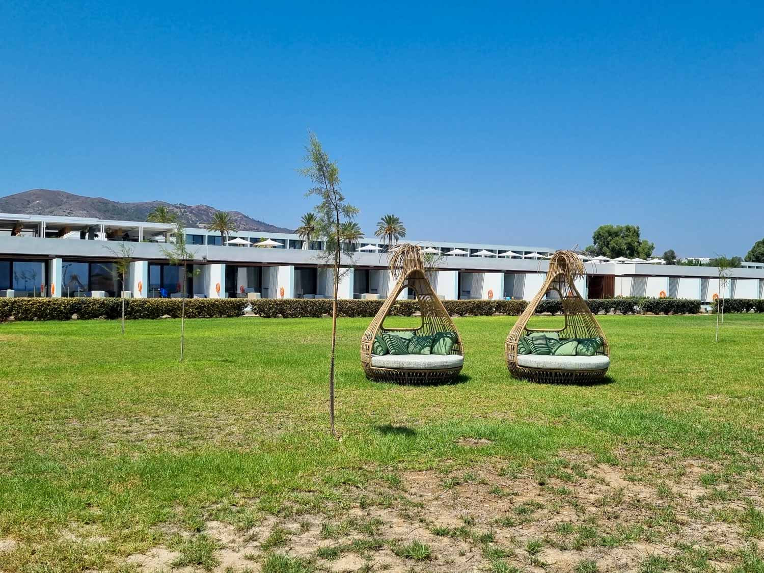 View from the beach to two comfy Nest seats on the grass with some of the rooms at Gennadi Grand Resort in the background and mountains behind - my Gennadi Grand Resort review of a stay in Rhodes with kids