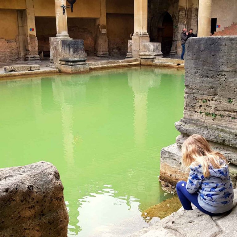 My daughter sits by the side of the water in the Roman Baths in Bath - the best known of my top things to do in Bath with kids