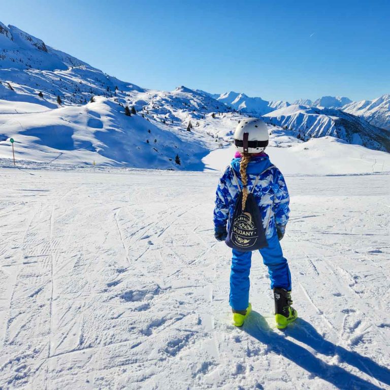 My daughter in colourful ski gear, ski boots and helmet, looks out to the snow covered mountains near Alpe d'Huez - my tips for a first family ski trip