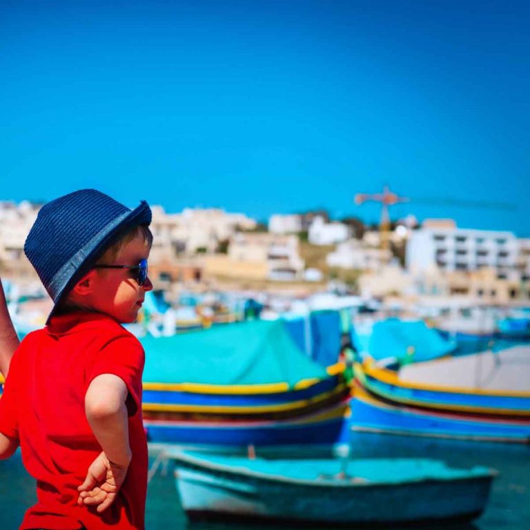 A boy and his mother in matching red clothes in front of a harbour in Malta with brightly coloured boats - why family holidays in Malta are a great idea