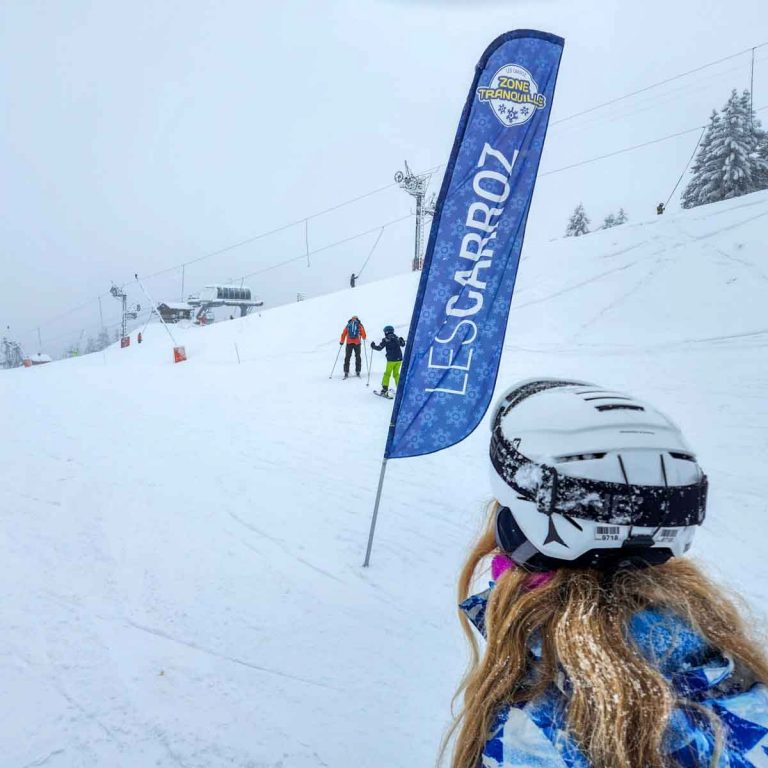 My daughter on the beginner green run at Les Carroz on a snowy day with a blue sign with the name of the resort - my tips for visiting Les Carroz with kids