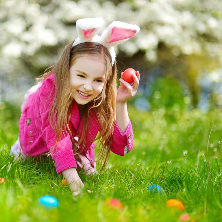 Girl wearing bunny ears and bright pink jacket hunting for bright eggs in the grass, with tree in blossom behind - the best Easter days out in London with kids