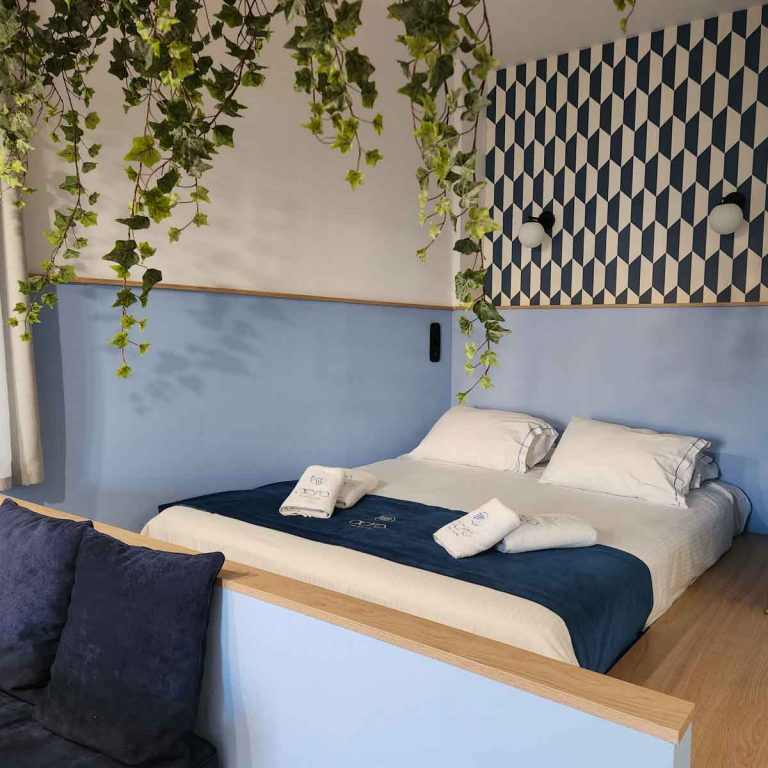 View of the double bed on its raised platform with artificial leaves and a blue and white geometric pattern above pale blue walls in our studio apartment at Acro Urban Suites aparthotel in Athens - my review of a stay with kids