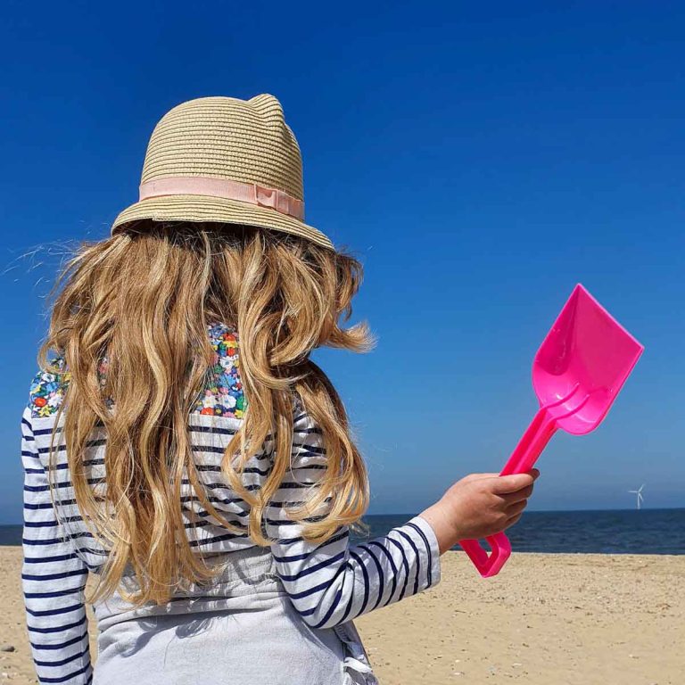 My daughter wearing a sunhat on a beach in Norfolk holding a bright pink spade - my pick of the best beaches in Norfolk with kids