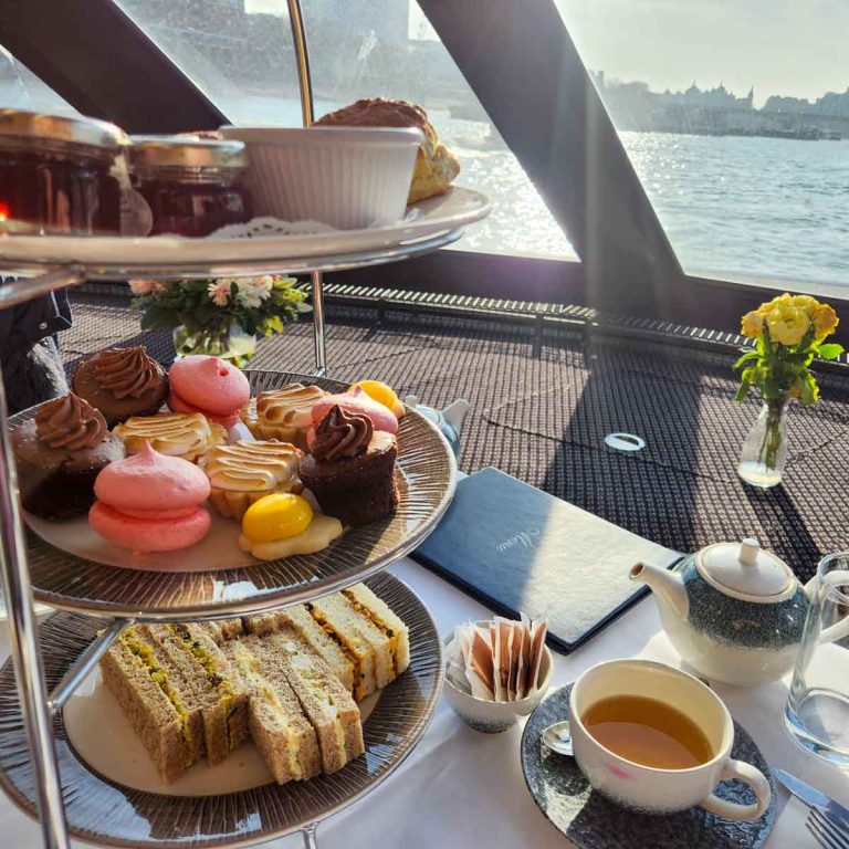 View of a three-tiered cake stand with sandwiches, cakes and scones and jam, teapot and tea cup on the table and the river Thames visible through the boat windows beyond - my London afternoon tea cruise review