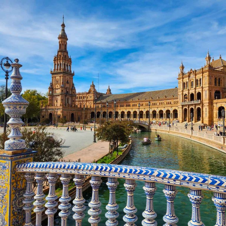 View across the blue, yellow and white tiled bridge to the canal and curving golden buildings of the Plaza de Espana - a visit is one of the unmissable things to do in Seville with kids