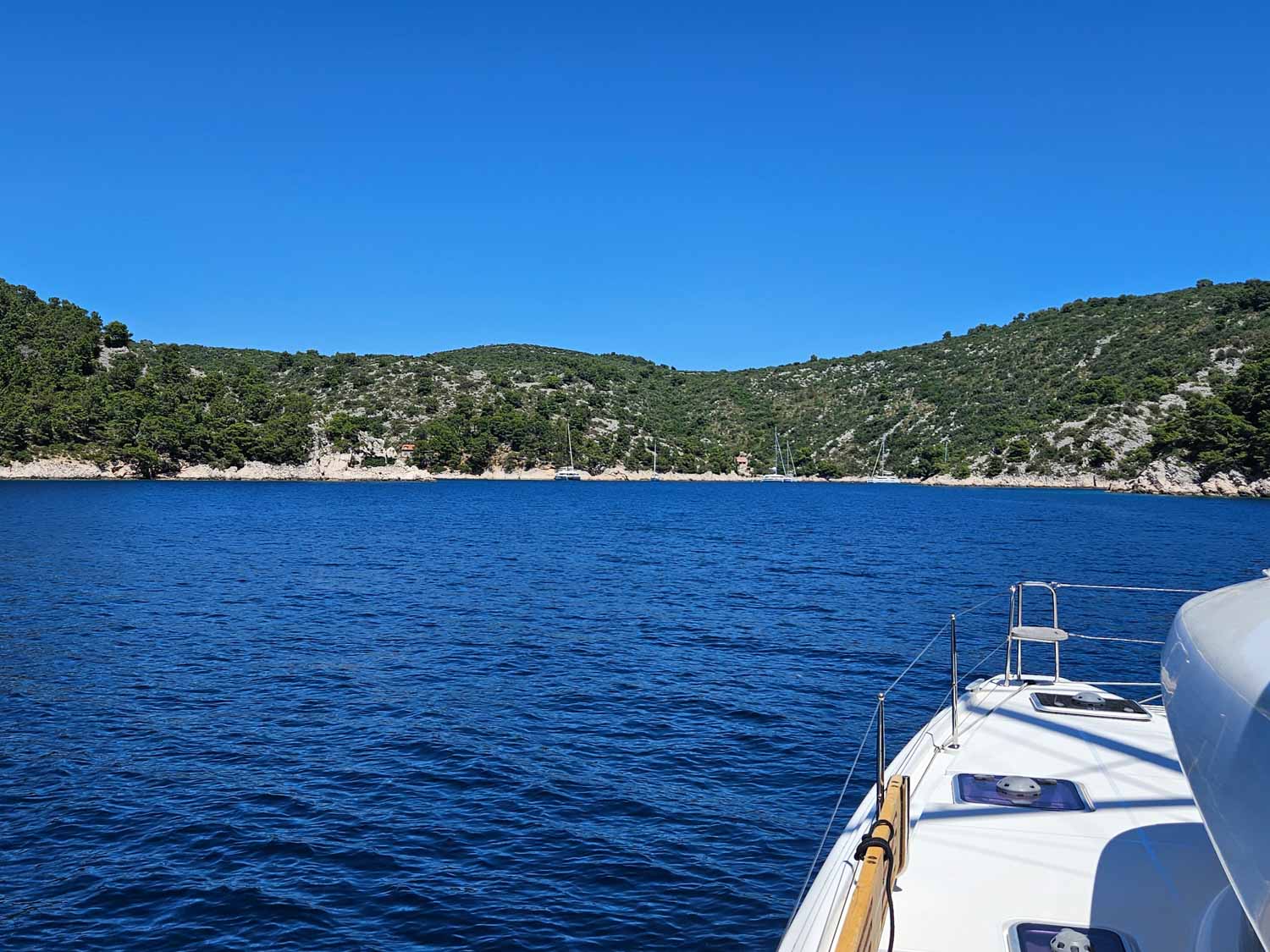 View across the front of our catamaran to deep blue water and a moored boat in front of the cliffs at a little bay on Solta, one of the stops on our Croatia sailing holiday with kids