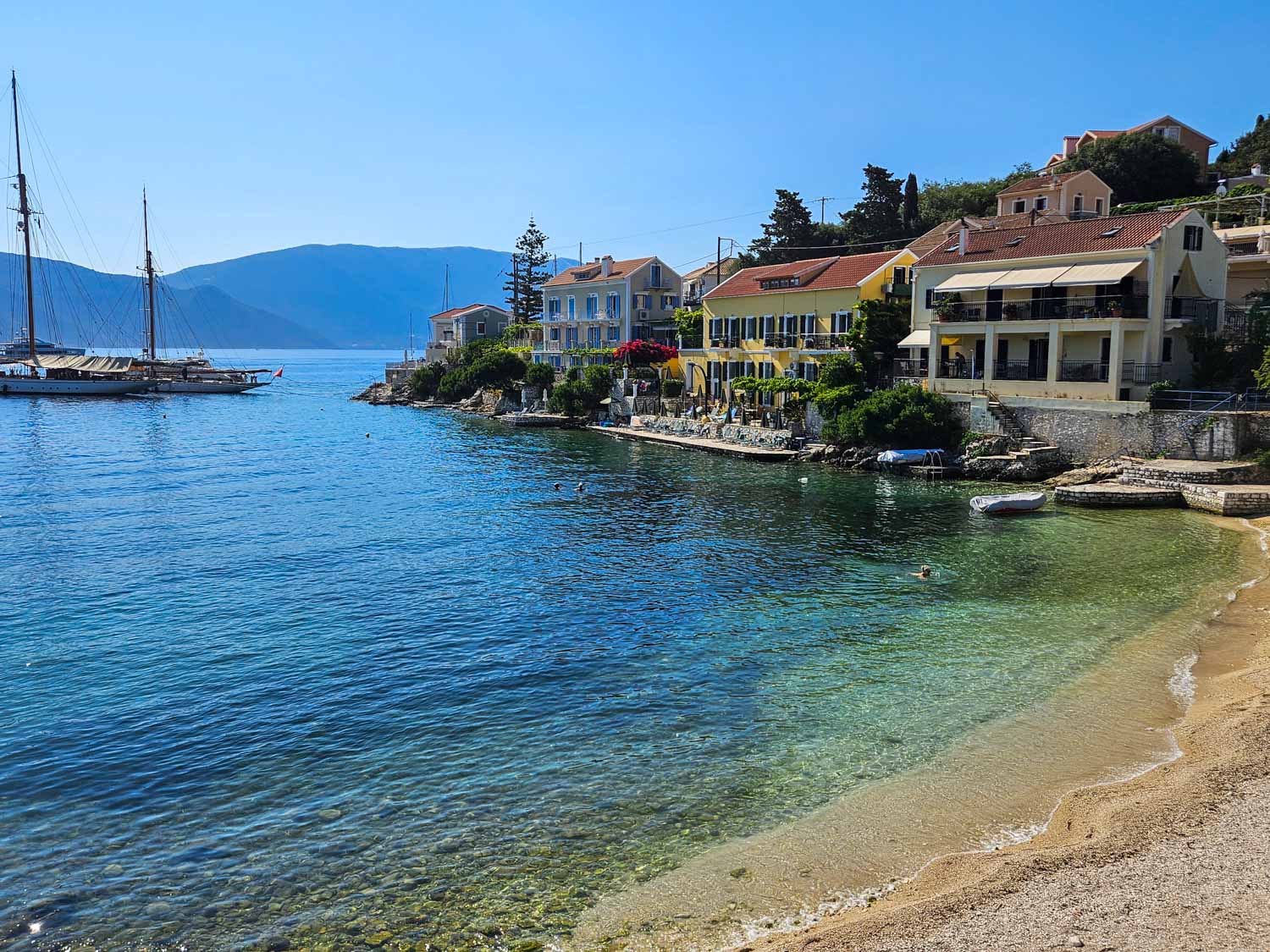 View of the little shingle beach with clear turquoise water by more colourful houses - what to do during one day in Fiskardo with kids