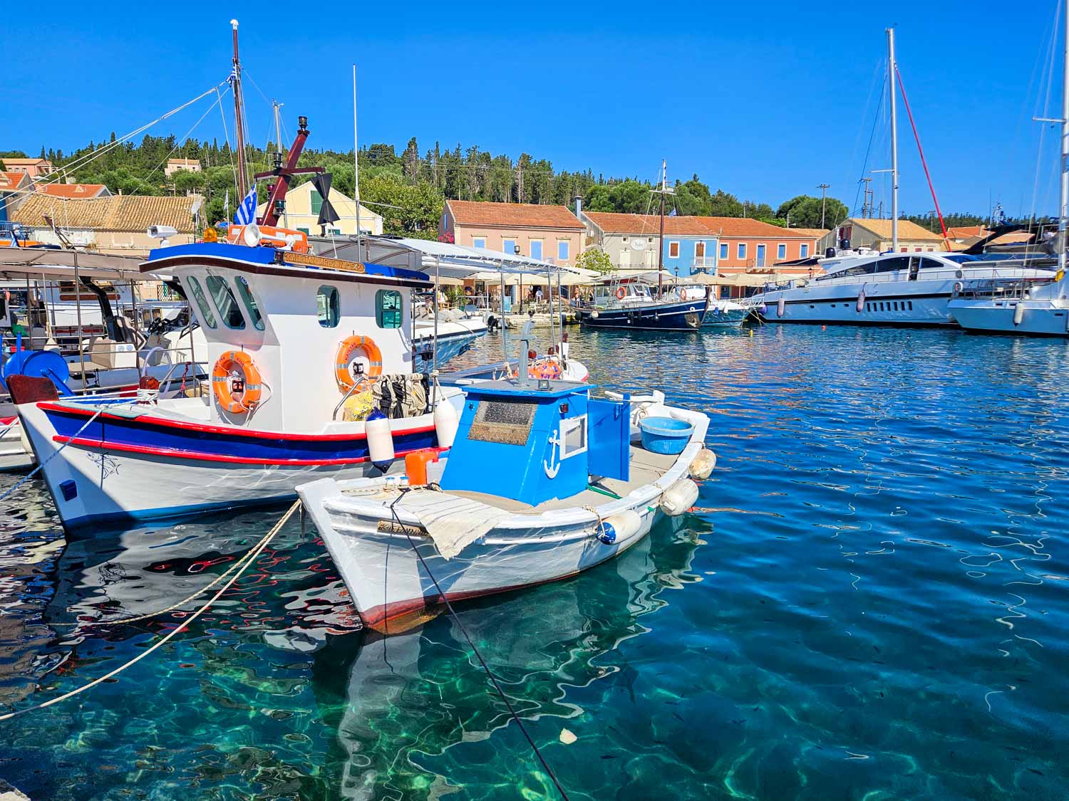View of the clear turquoise water in the harbour with colourful buildings behind and boats in the foreground - what to do during one day in Fiskardo with kids