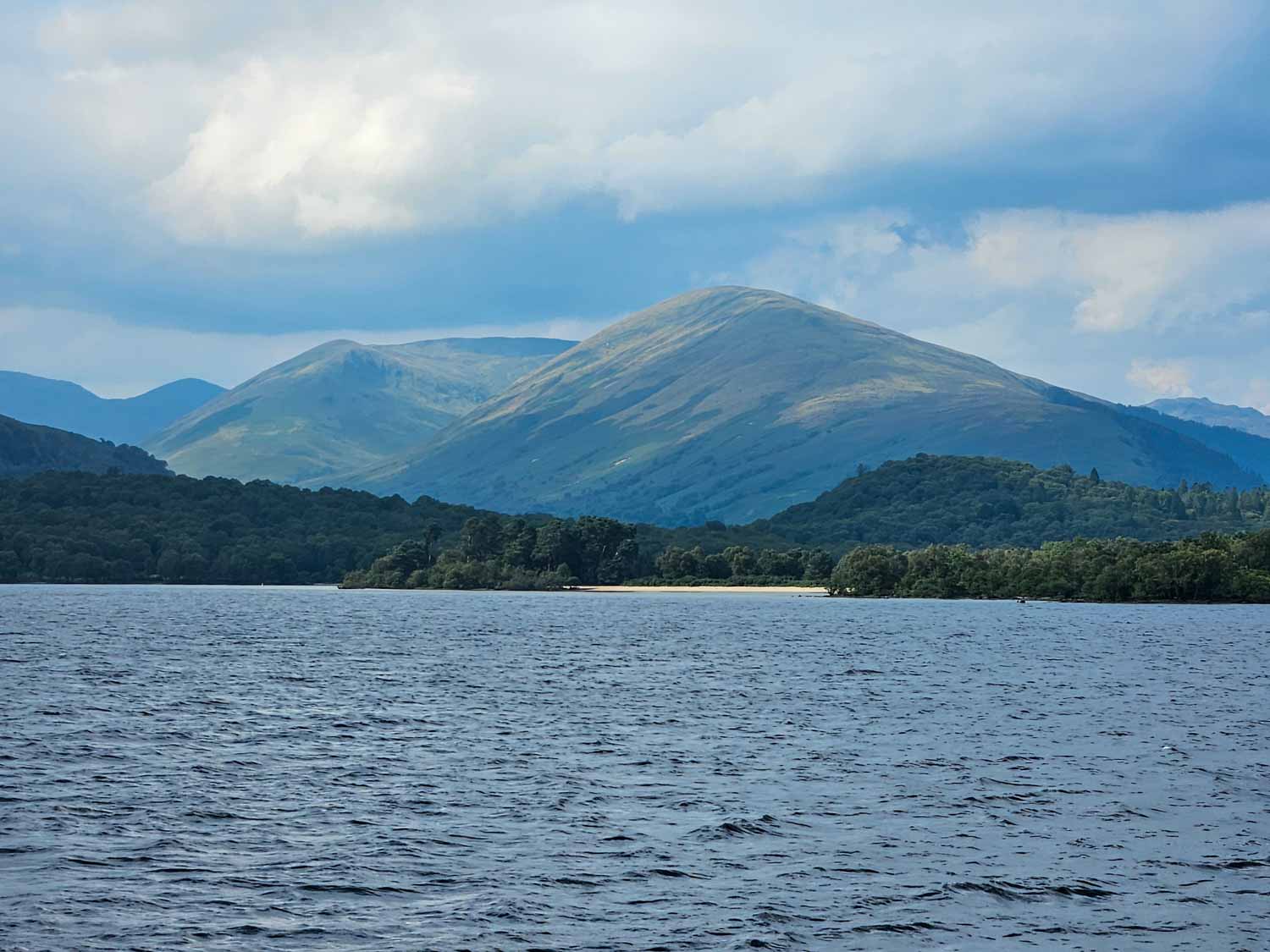 View across the water to a small golden beach surrounded by trees and hills in the background - a boat trip is one of the best things to do in Loch Lomond with kids
