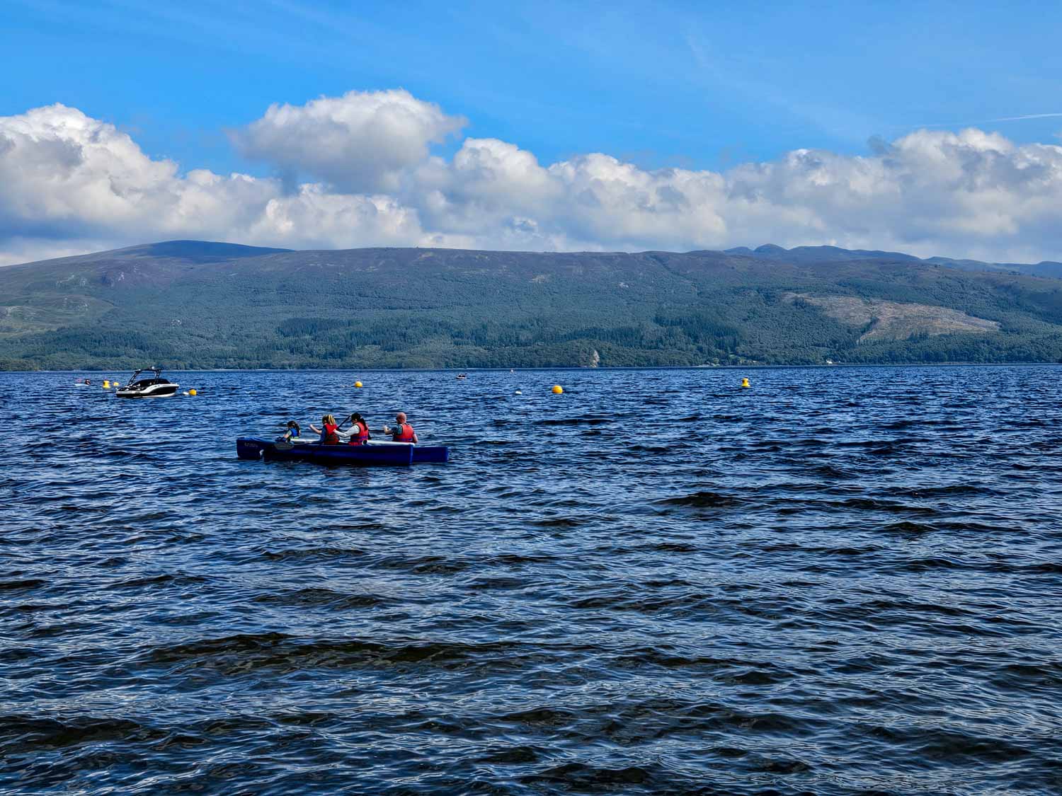View of a group in a canoy and small boat just seen on the blue waters of the loch at Luss - my pick of the best things to do in Loch Lomond with kids